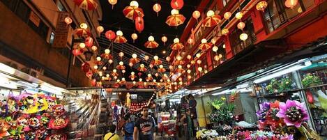 A market with stalls selling flowers and other goods, under a canopy of hanging lanterns.