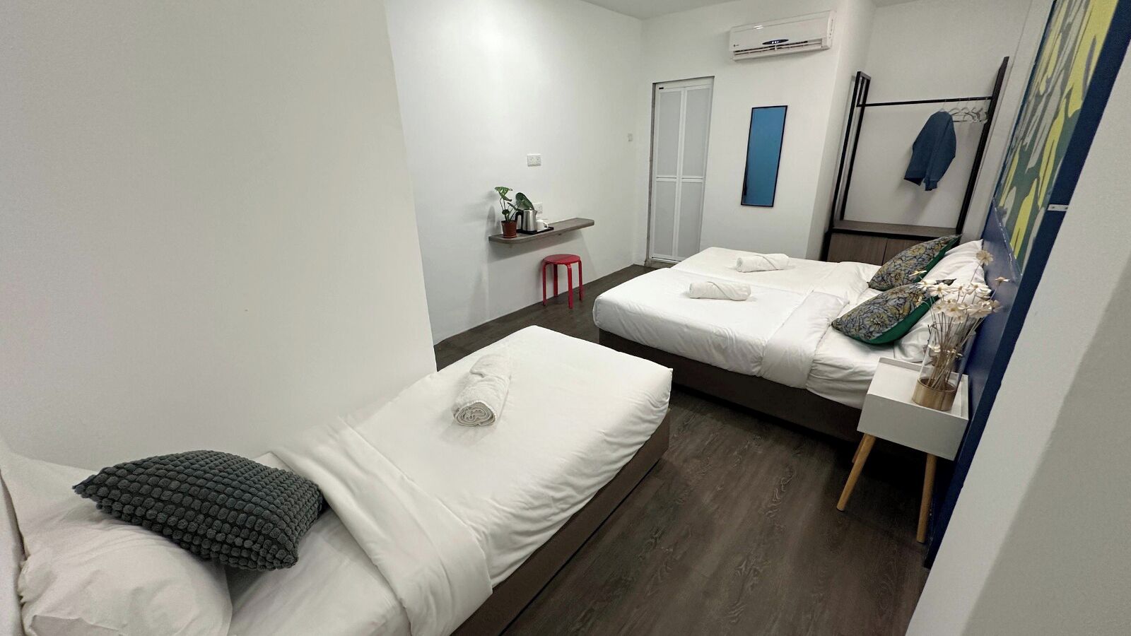 A hotel room with two single beds, a small table with a vase of flowers, a red stool, a wardrobe, and a mirror on the wall.