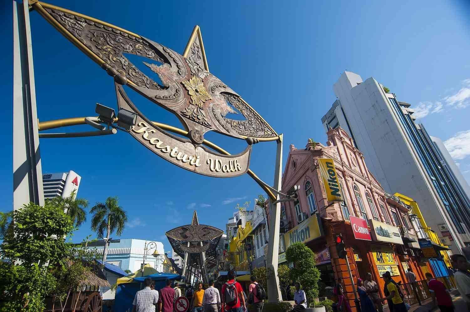 A large, ornate metal sign with the inscription "Kasturi Wall" in a bustling urban street with modern buildings and shops.