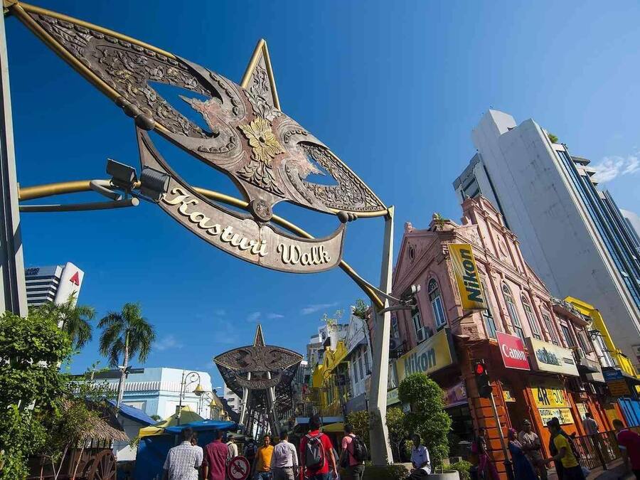 A large, ornate metal sign with the inscription "Kasturi Wall" in a bustling urban street with modern buildings and shops.