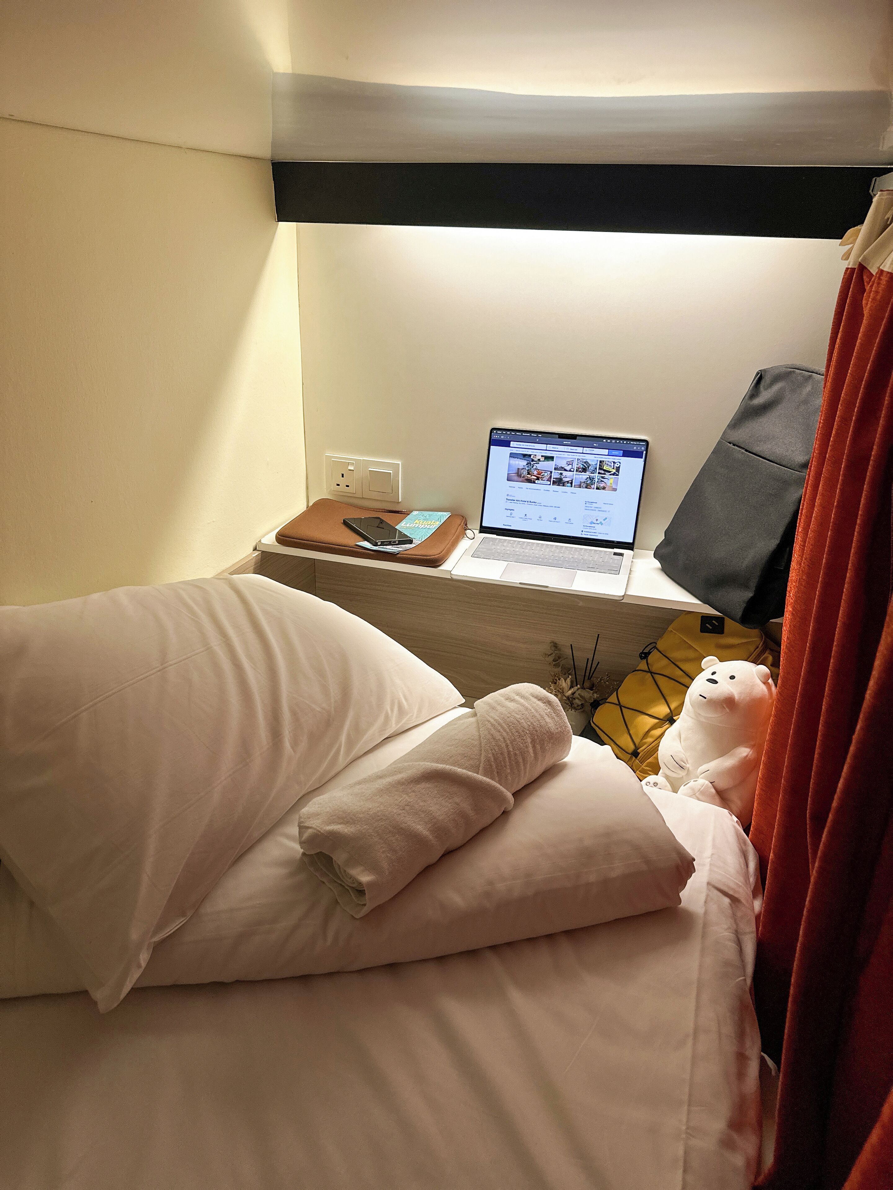 A neatly made bed with pillows, a laptop on a bedside table, a smartphone, and a teddy bear.