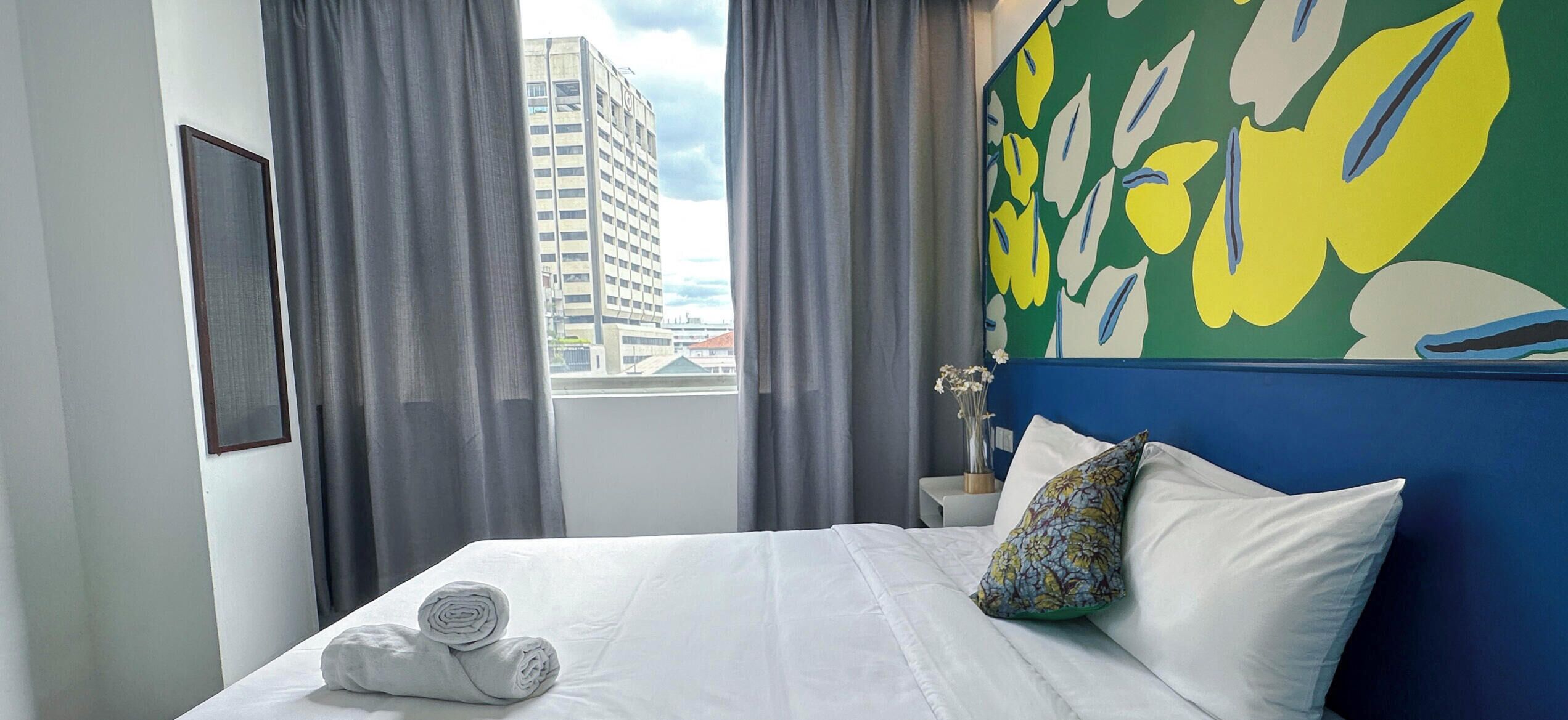 Habitación de hotel con cama, ropa de cama blanca, cabecera azul, almohada estampada con flores, mesita de noche y vista de un edificio a través de la ventana.