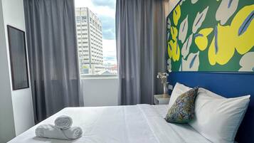 Habitación de hotel con cama, ropa de cama blanca, cabecera azul, almohada estampada con flores, mesita de noche y vista de un edificio a través de la ventana.
