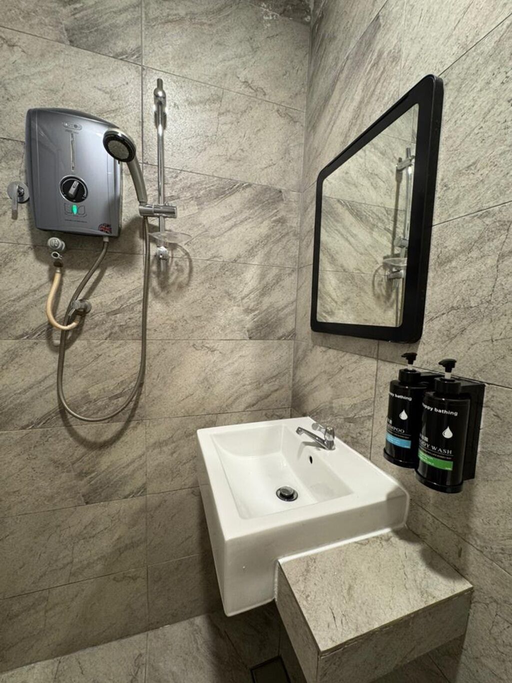 Un baño con un lavamanos blanco, un cabezal de ducha de mano, un espejo montado en la pared y dos botellas de champú y gel de baño.