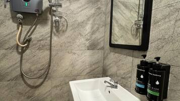 Un baño con un lavamanos blanco, un cabezal de ducha de mano, un espejo montado en la pared y dos botellas de champú y gel de baño.