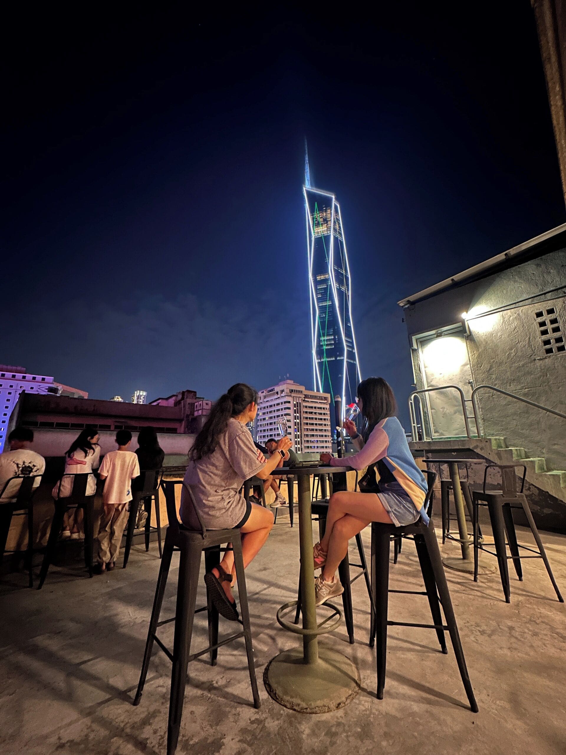 An outdoor seating area with tables and chairs, a view of a tall building with illuminated windows, and a staircase leading to an upper level.