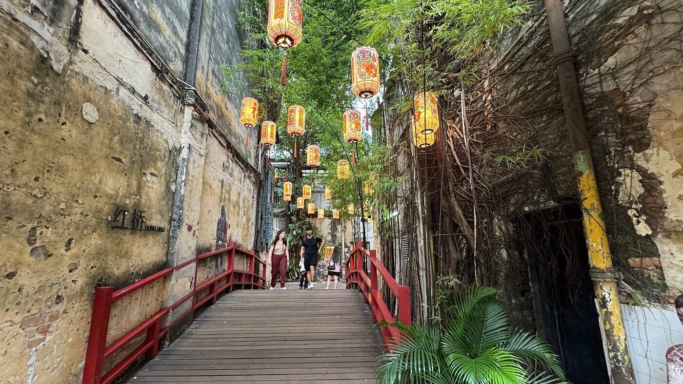 A narrow alleyway with a wooden bridge, red railings, and hanging lanterns.