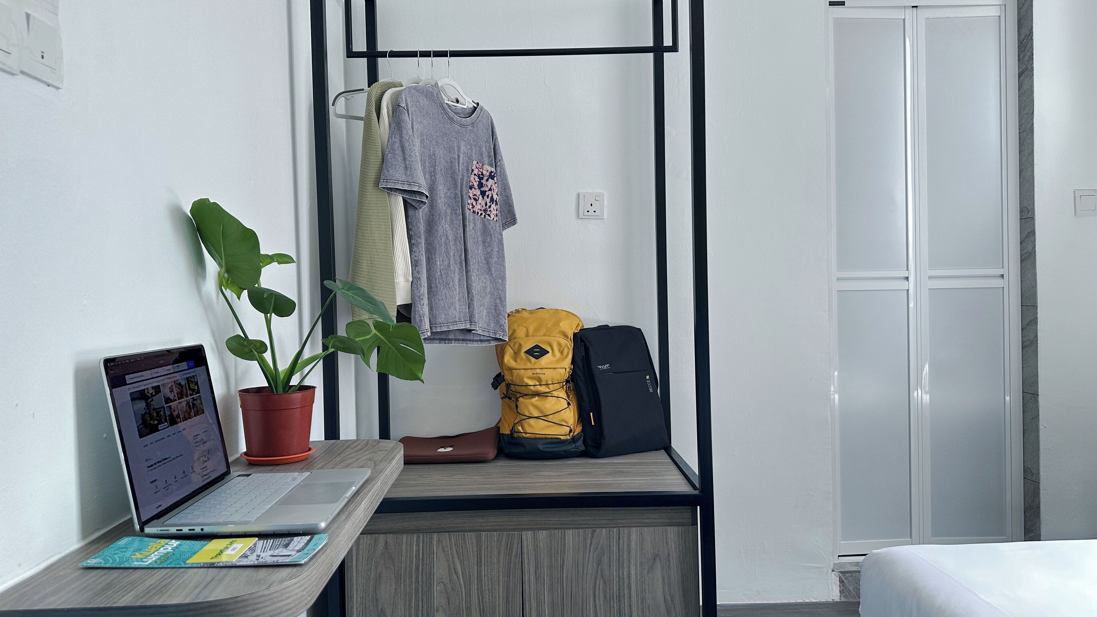 Ein kleines Zimmer mit einem Holztisch, einem Laptop, einer Topfpflanze, einem gelben Rucksack und einer grauen Hoodie, die auf einem Gestell hängt.