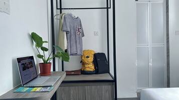 Ein kleines Zimmer mit einem Holztisch, einem Laptop, einer Topfpflanze, einem gelben Rucksack und einer grauen Hoodie, die auf einem Gestell hängt.