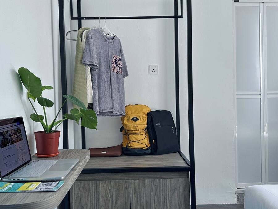 Ein kleines Zimmer mit einem Holztisch, einem Laptop, einer Topfpflanze, einem gelben Rucksack und einer grauen Hoodie, die auf einem Gestell hängt.