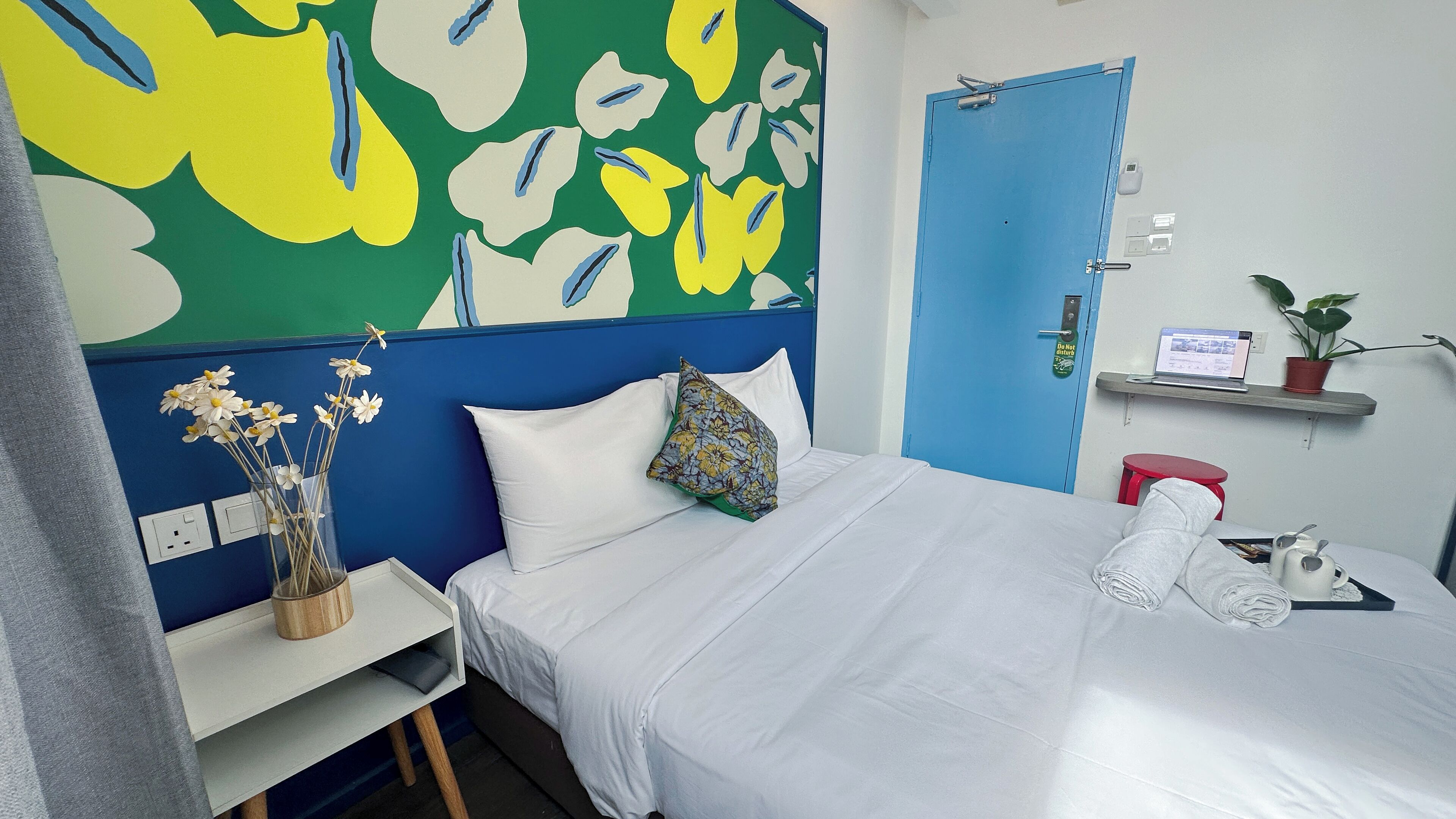 A neatly made bed with white linens, a blue door, a small desk with a vase of flowers, and a laptop on a shelf.