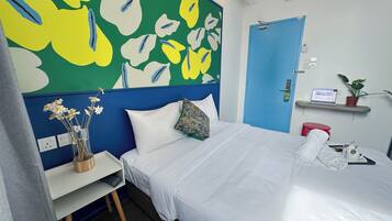 A neatly made bed with white linens, a blue door, a small desk with a vase of flowers, and a laptop on a shelf.