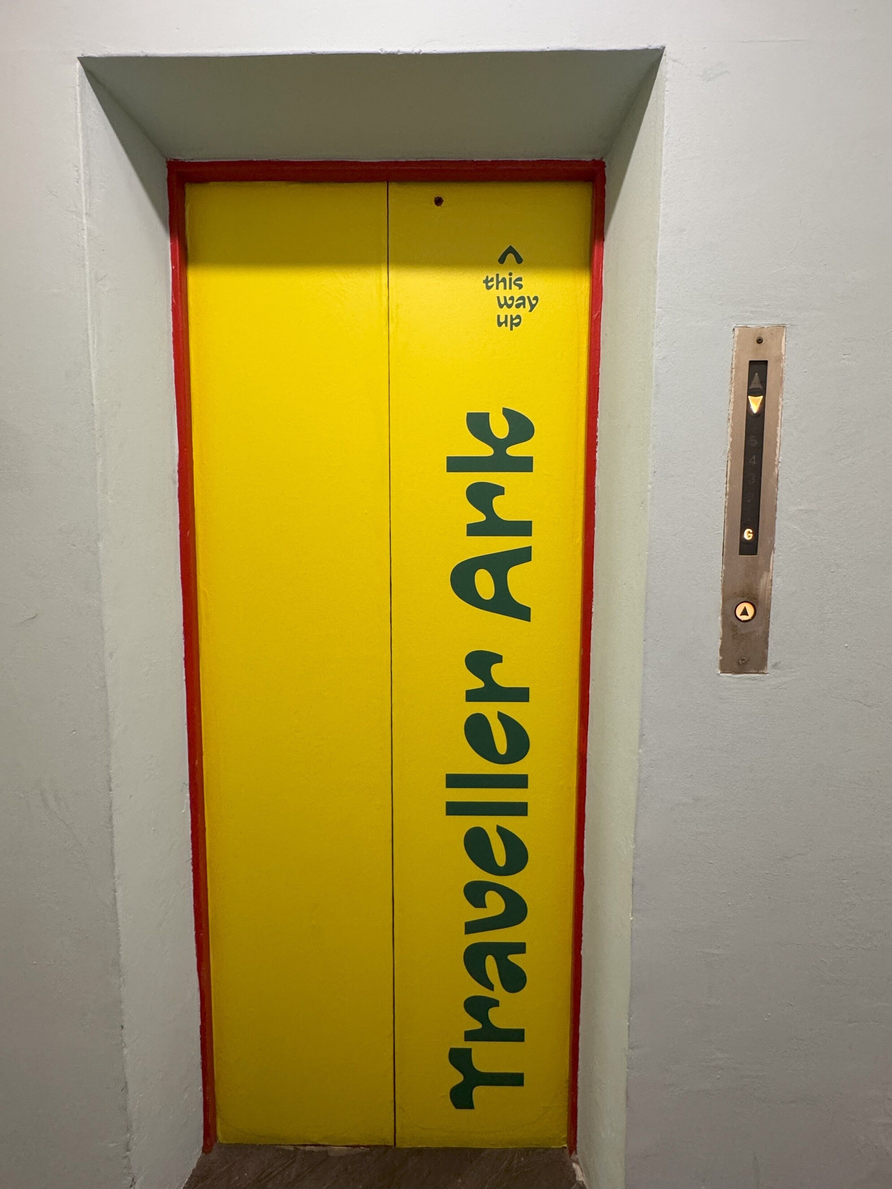 Un ascensor amarillo con la leyenda 'Traveller Ark', un borde rojo y un pequeño letrero sobre la puerta que dice 'subir'.
