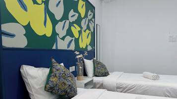 Habitación de hotel con dos camas, ropa de cama blanca, un florero con flores y un cuadro abstracto colorido en la pared.