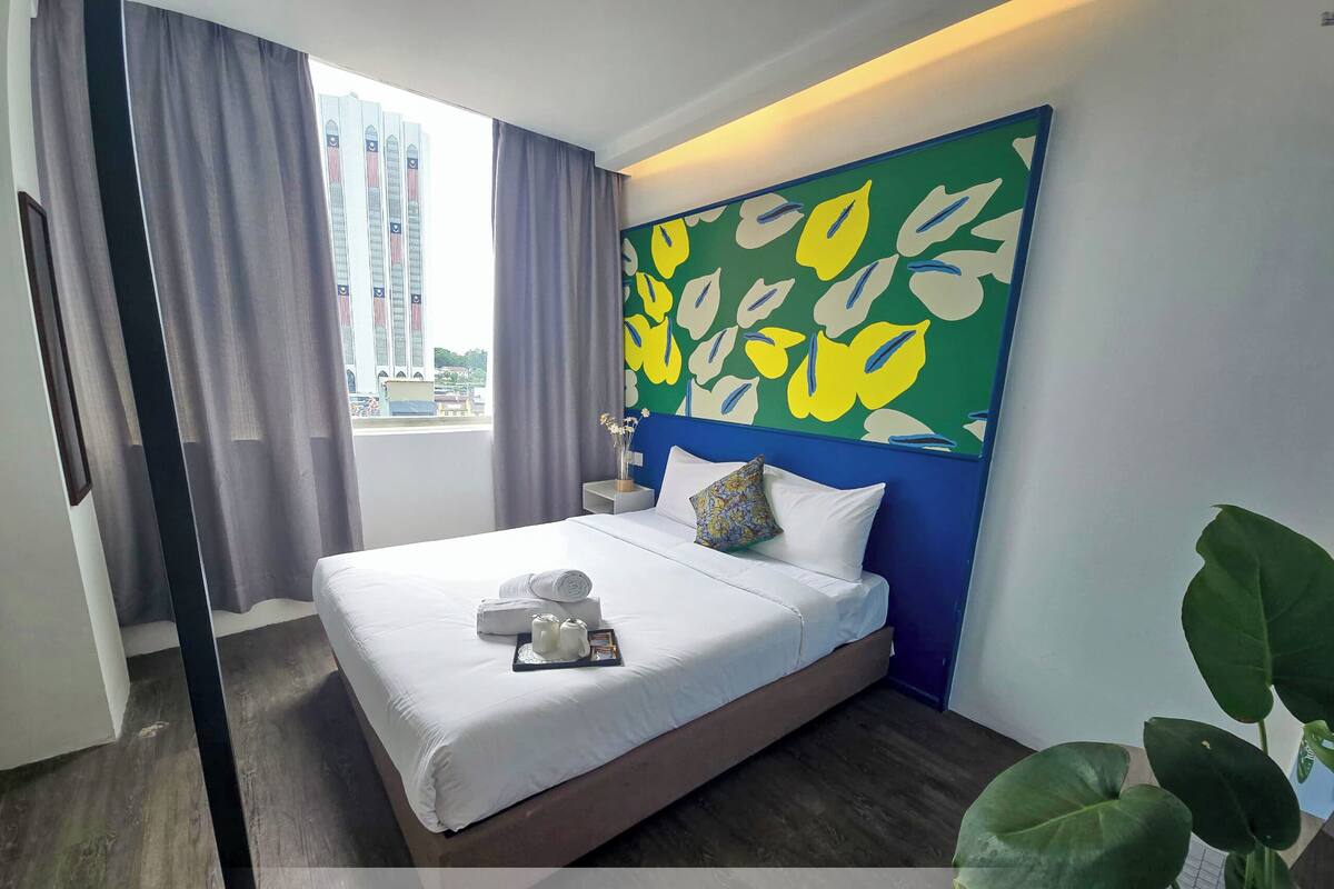 A hotel room with a bed, a headboard with a leaf pattern, a window with curtains, and a view of buildings outside.