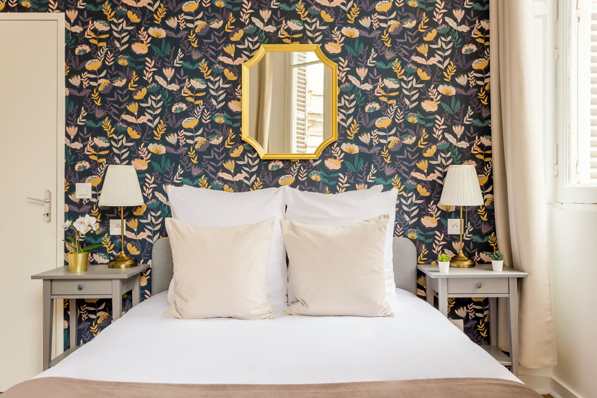 Ein Schlafzimmer mit floralem Wandpapier, einem großen Bett mit weißen Bettwäsche, zwei Nachttischen mit Lampen und einem Spiegel über dem Bett.