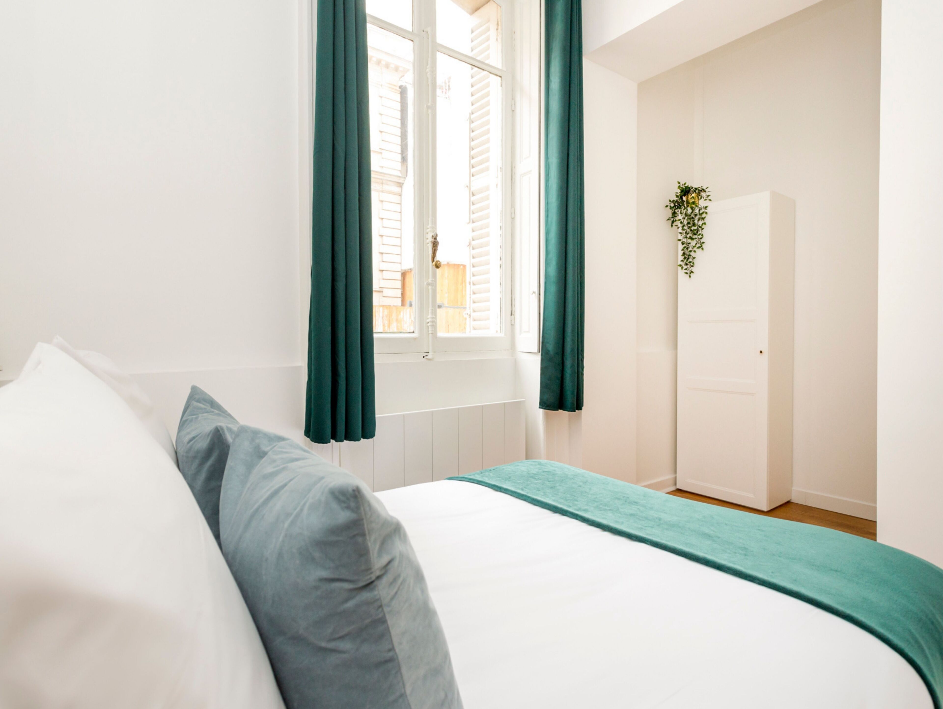 Een slaapkamer met een bed, witte muren, een raam met groene gordijnen en een deur.