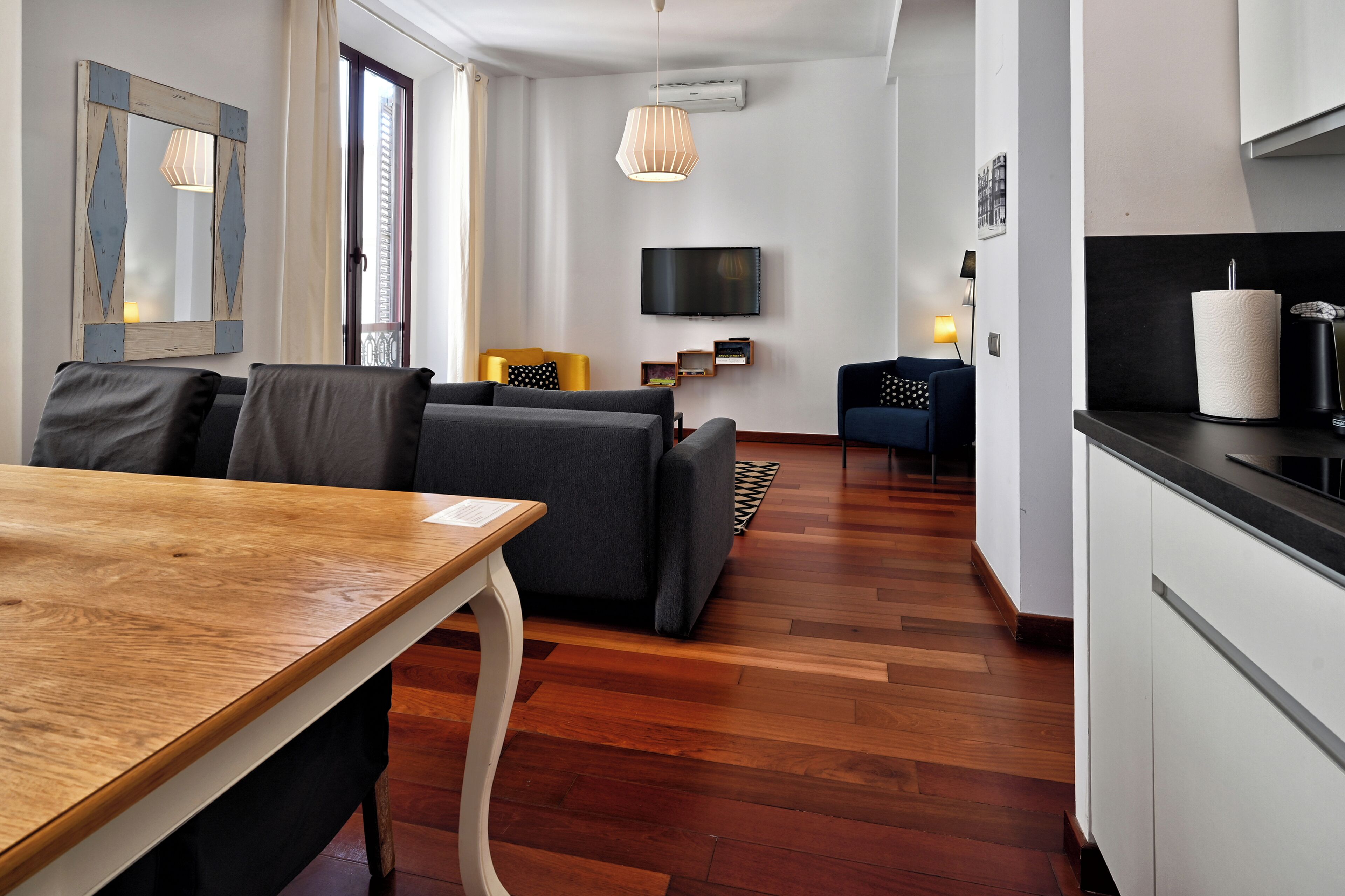 A modern living room with a wooden dining table, grey sofas, a TV, and a kitchen area with a black countertop.