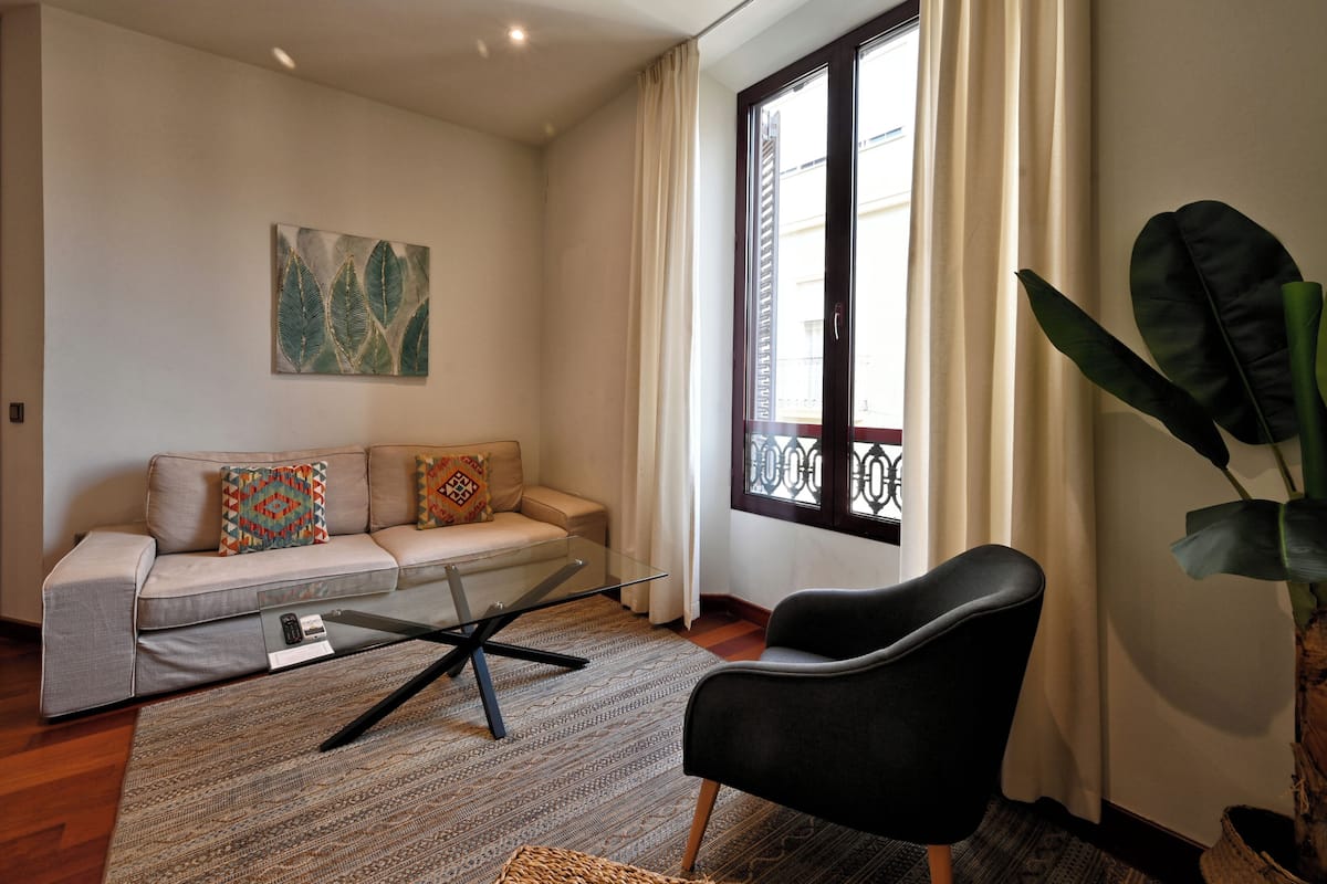 Ein Wohnzimmer mit einer Couch, einem schwarzen Sessel, einem Glastisch und einer großen Pflanze.