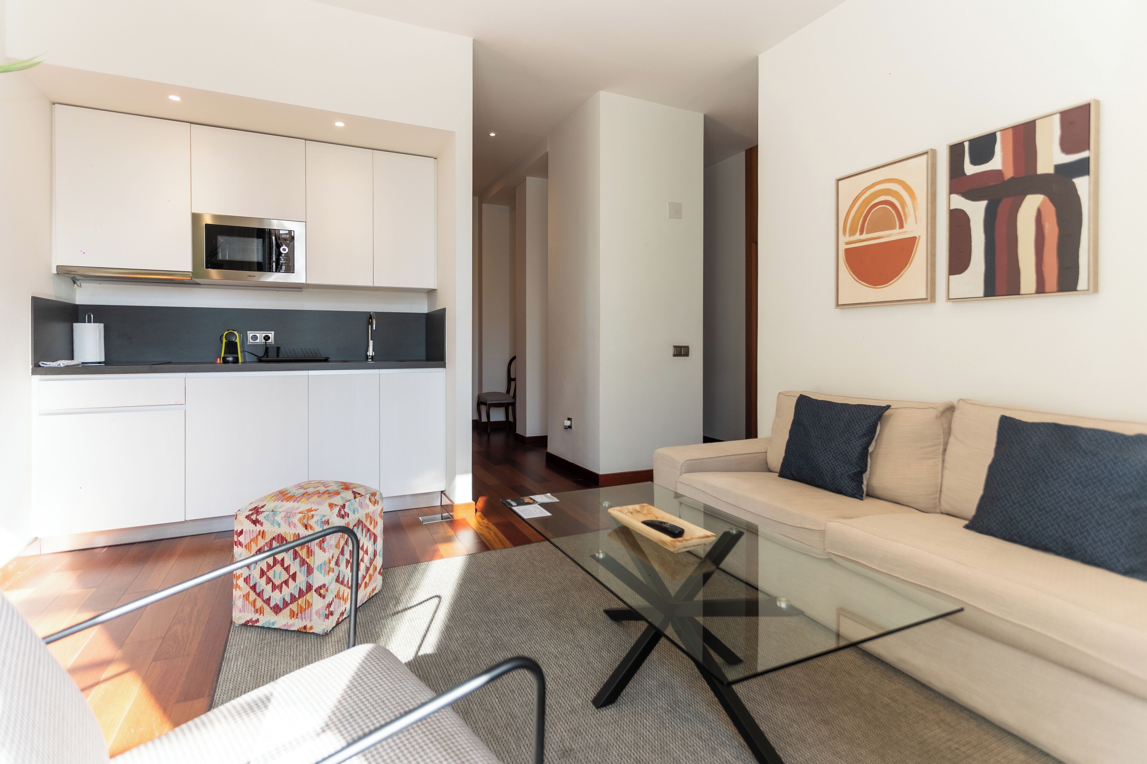 A modern living room with a glass coffee table, a sofa, and a kitchen area with white cabinets and a microwave.