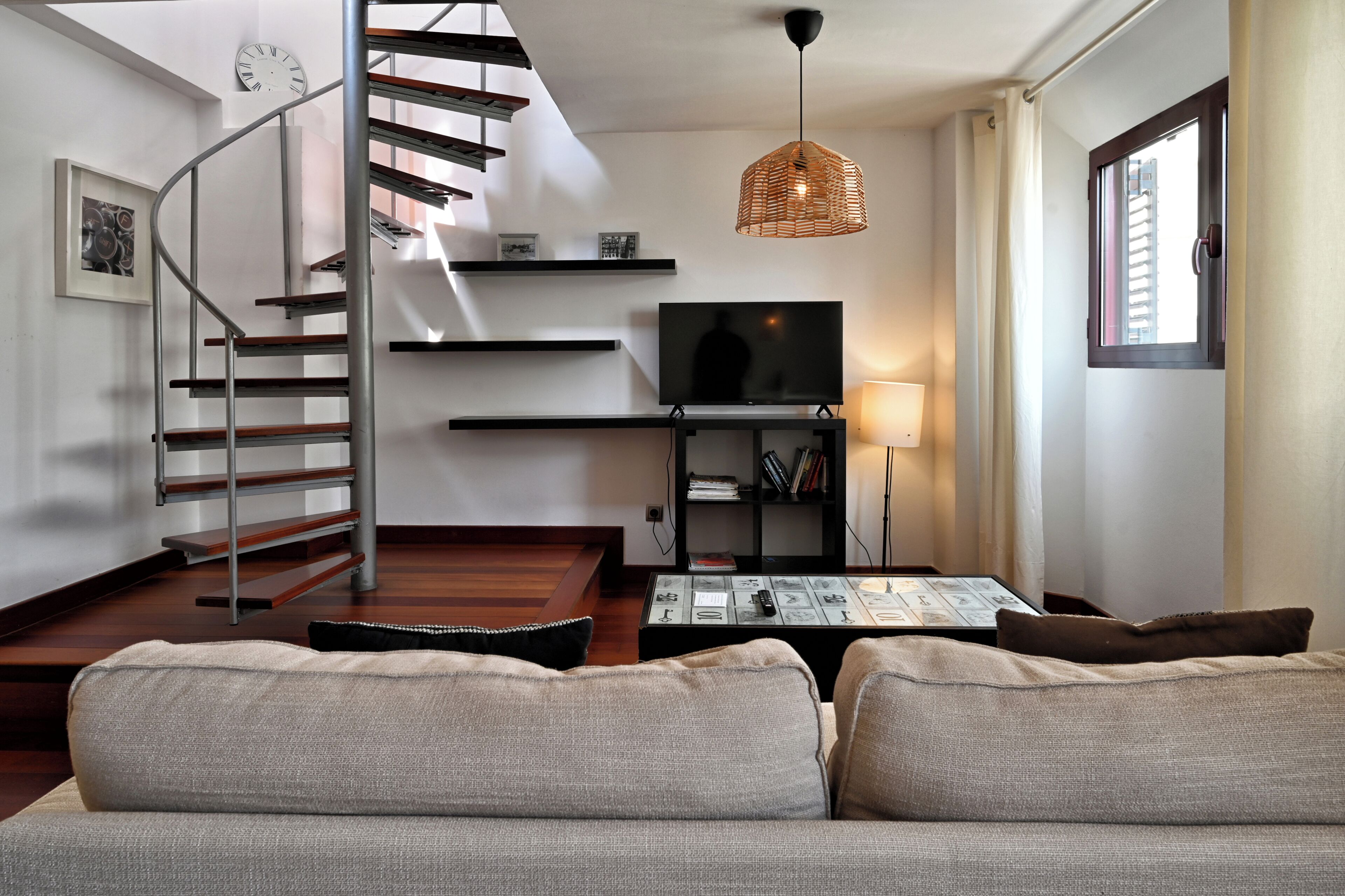A modern living room with a staircase, a flat-screen TV, a coffee table, and a bookshelf.