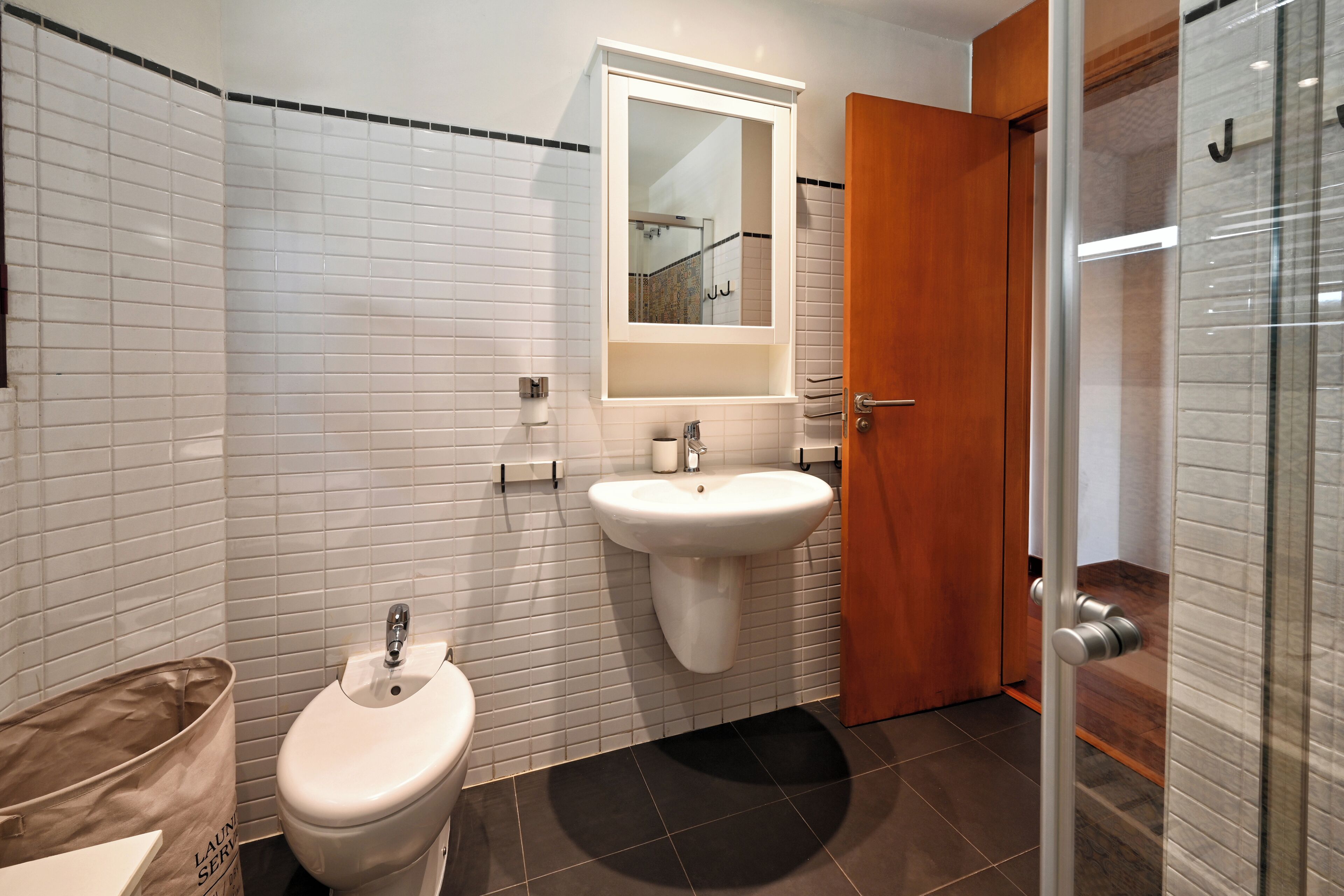 Un baño con paredes de azulejos blancos, un lavamanos blanco, un inodoro y una puerta de madera.