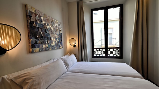 Ein Hotelzimmer mit zwei Betten, einem geometrischen Kunstwerk an der Wand, einem Fenster mit Blick auf einen Balkon und einer Wandleuchte.