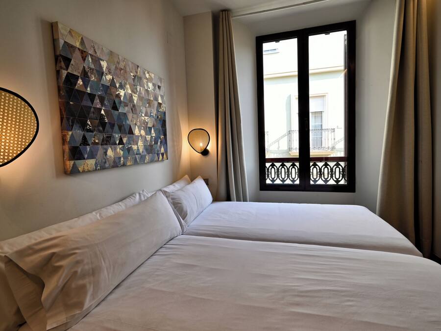 Una habitación de hotel con dos camas, una obra de arte geométrica en la pared, una ventana con vistas a un balcón y una lámpara fijada a la pared.