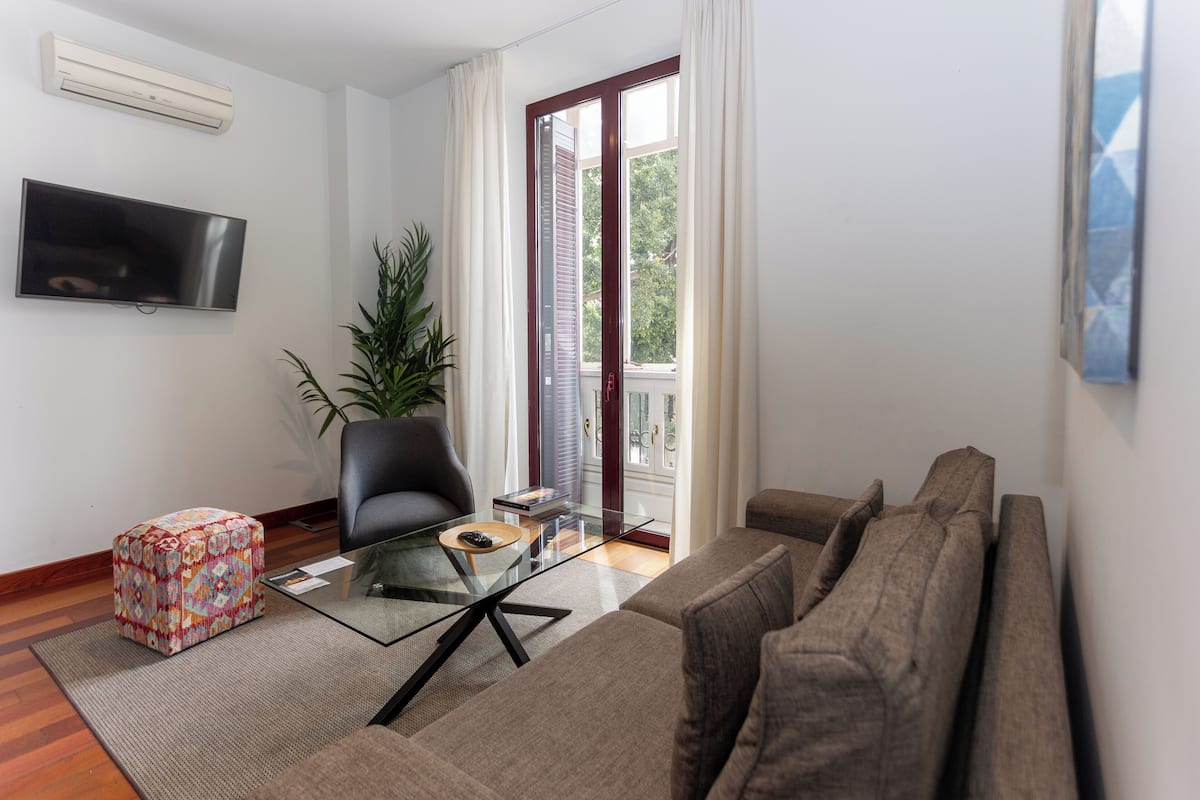 Ein modernes Wohnzimmer mit einem Glastisch, einer Couch, einem Sessel und einem an der Wand befestigten Fernseher.