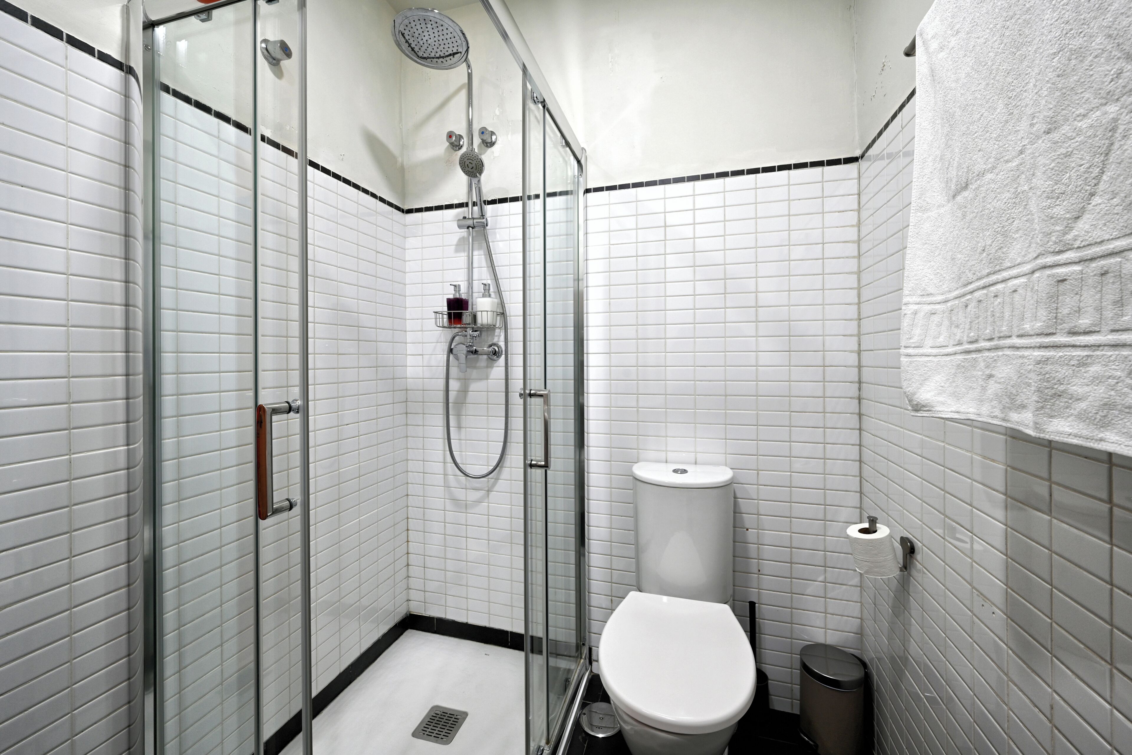 Et badeværelse med en glasvæg omkring brusenichen, et hvidt toilet og en skraldespand.