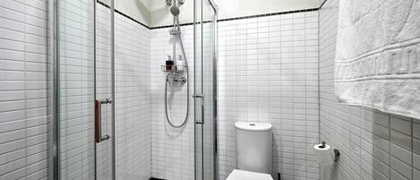 Et badeværelse med en glasvæg omkring brusenichen, et hvidt toilet og en skraldespand.