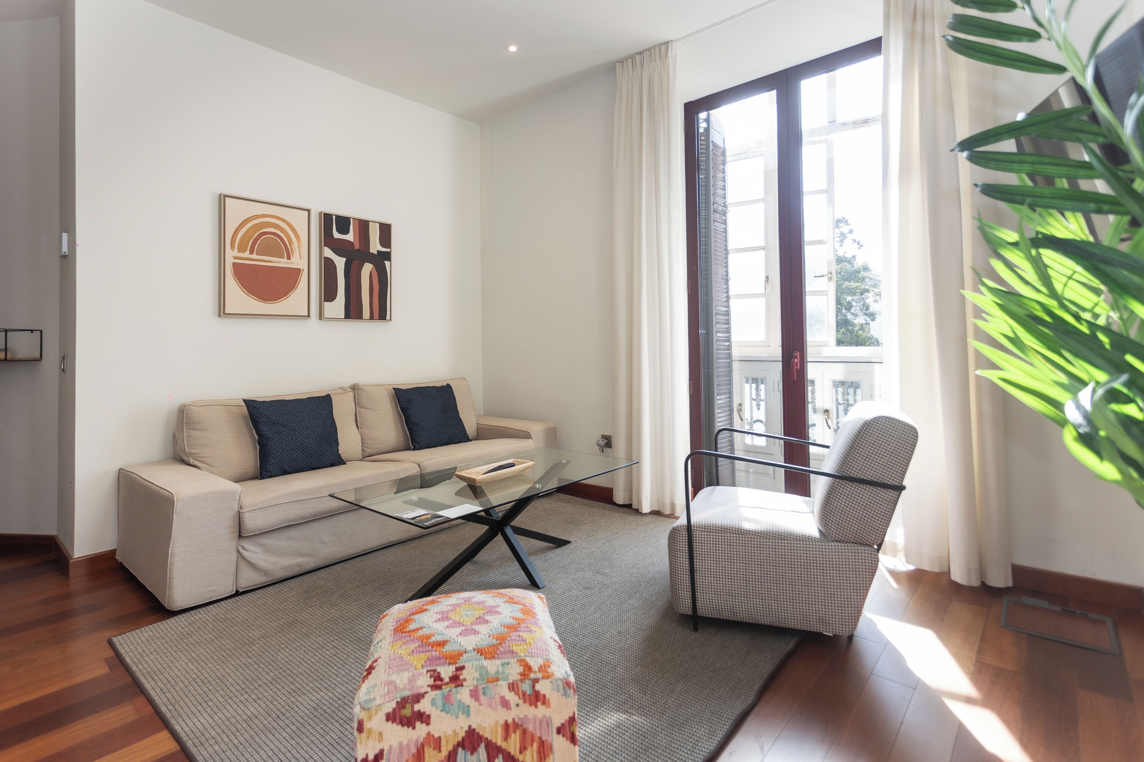 A modern living room with a sofa, a glass coffee table, a patterned ottoman, a chair, and artwork on the wall.