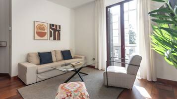 A modern living room with a sofa, a glass coffee table, a patterned ottoman, a chair, and artwork on the wall.