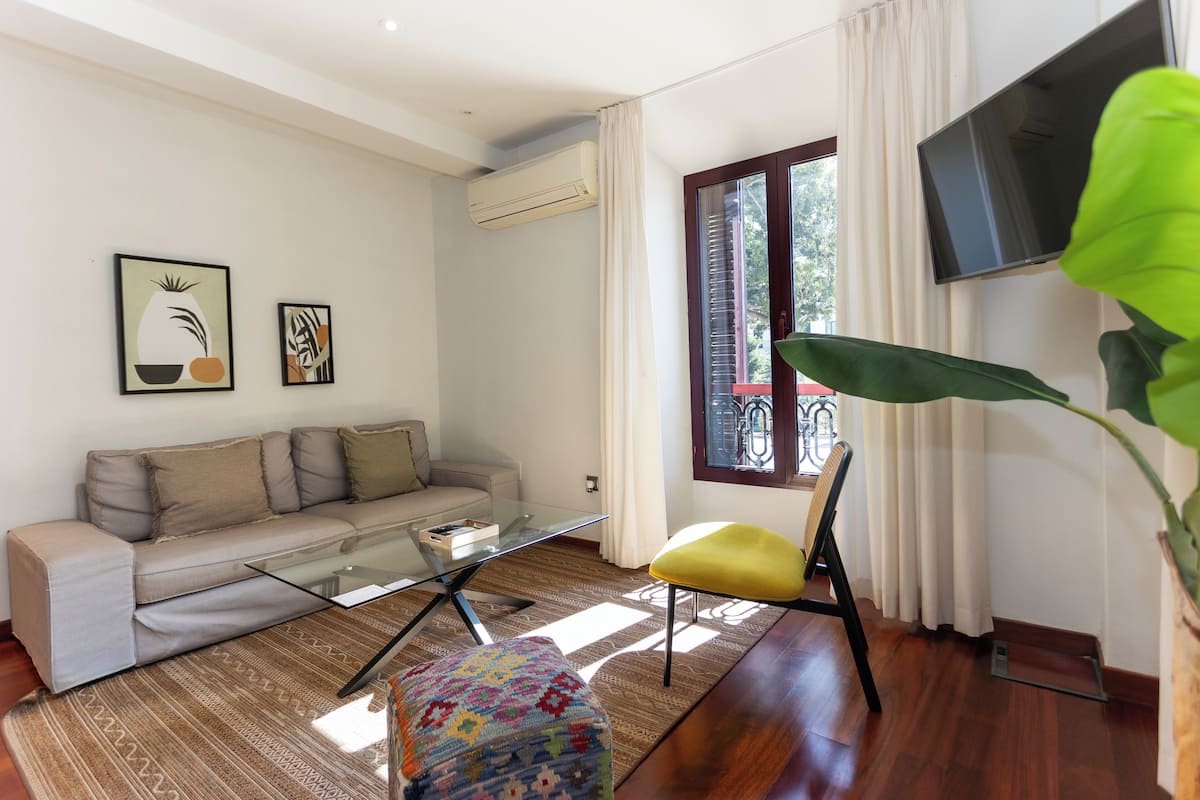 Ein Wohnzimmer mit einer Couch, einem Glastisch, einem gelben Sessel, einer großen Grünpflanze und einem an der Wand montierten Fernseher.