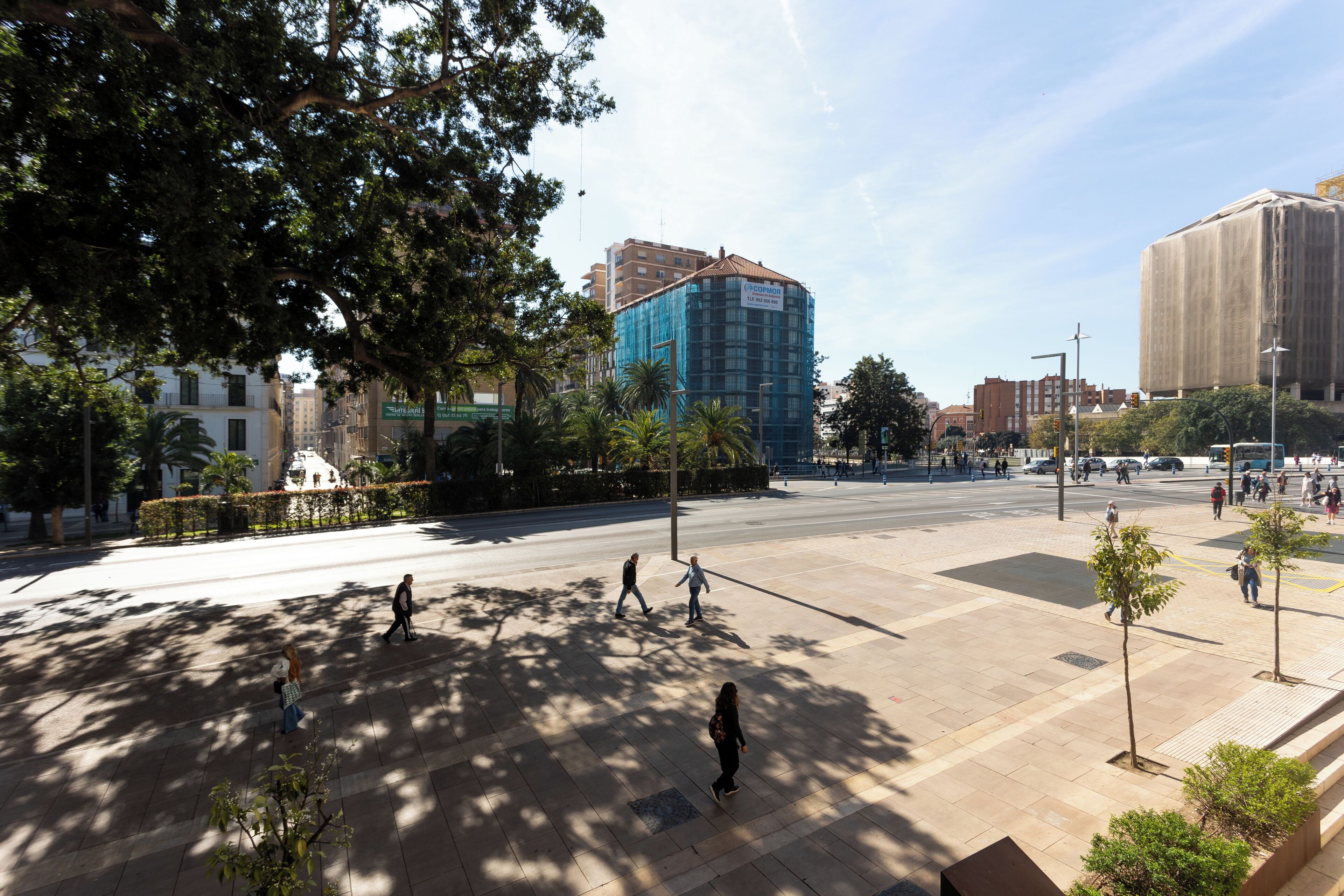 A spacious urban square with trees, a building with a blue facade, and a large, modern structure in the background.