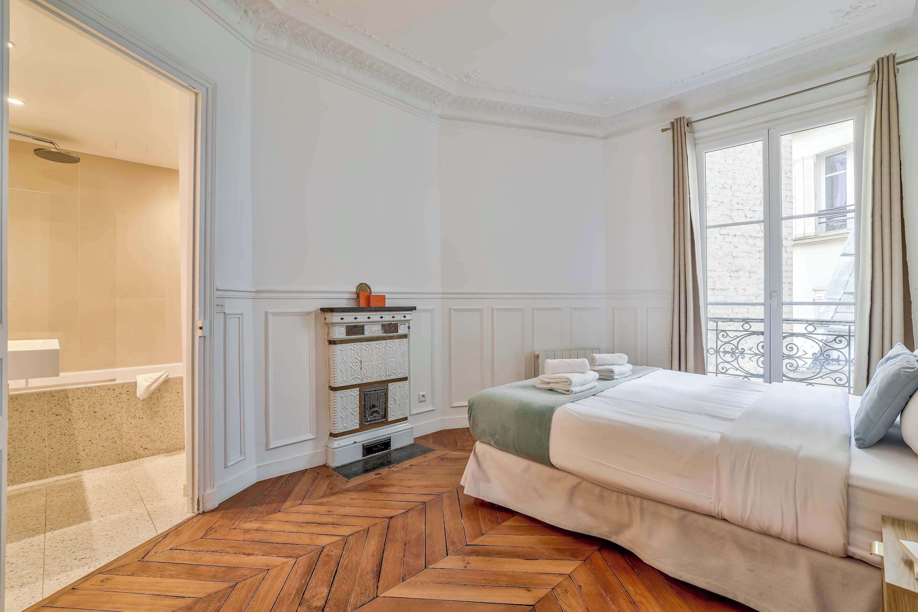 A bedroom with a herringbone floor, a bed with a green and white duvet, a fireplace, and a window with curtains.