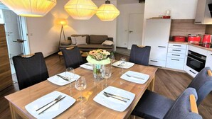 A dining area with a wooden table set for six, surrounded by chairs, with a sofa and a kitchen in the background. - Moderne Ferienwohnung in Winterberg (Winterberg)