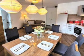 Un'area pranzo con un tavolo in legno apparecchiato per sei persone, circondato da sedie, con un divano e una cucina sullo sfondo.