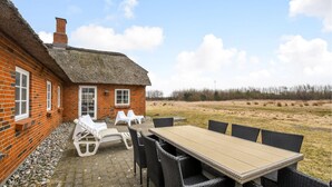 Ein Backsteinhaus mit einem Strohdach, eine Terrasse mit Gartenmöbeln und Blick auf ein Wiesenfeld.