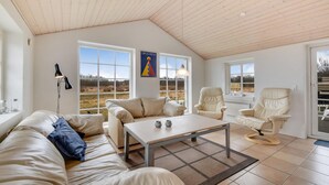 Ein geräumiges Wohnzimmer mit hohen Decken, großen Fenstern mit Blick auf eine Wiese, einem Sofa, einem Couchtisch und zwei Sesseln.