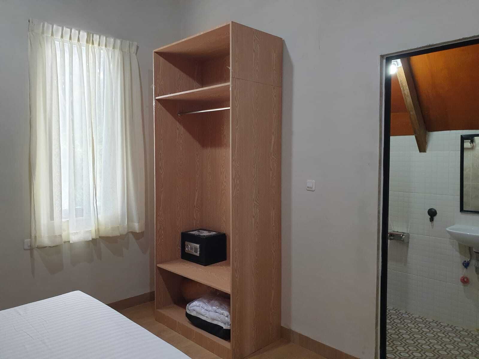 A room with a wooden wardrobe, a bed, a bathroom with a sink and mirror, and a window with curtains.
