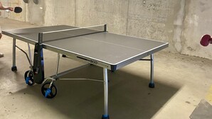 Une table de ping-pong portative, munie de roulettes, portant la mention « 900 Outdoor 2 », dans une pièce en béton où l’on peut voir des tuyaux et un mur rugueux.