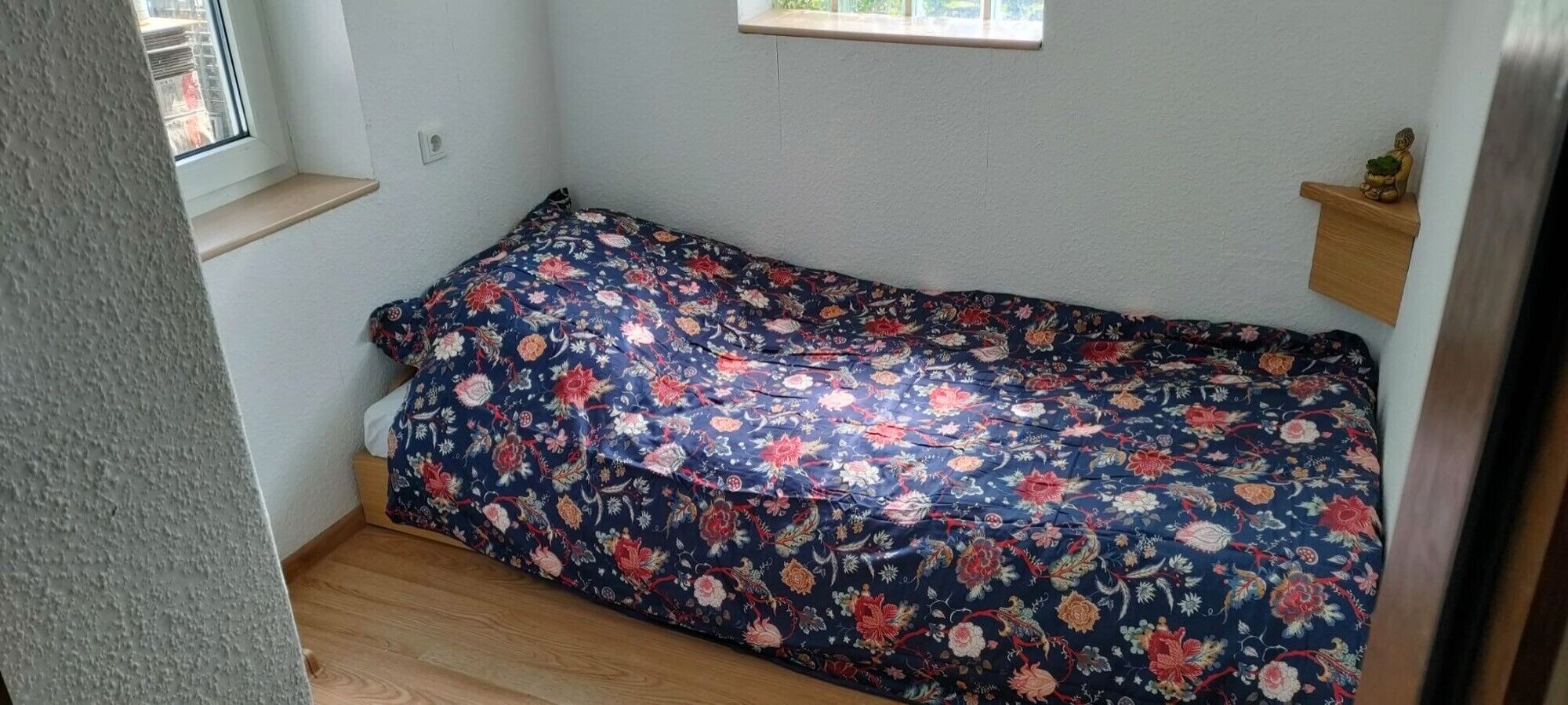 Ein kleines Zimmer mit einem blumengedeckten Bett, einem Fenster und einem kleinen Regal mit einer Buddha-Statue.