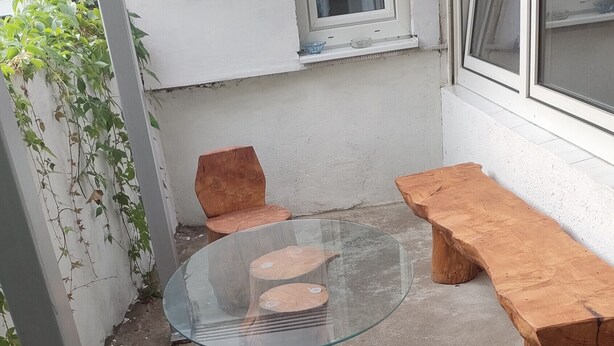 Une table à plateau en verre, entourée de tabourets et de bancs en bois, est placée sur une terrasse en béton. Un mur blanc et des fenêtres se dressent en arrière-plan.