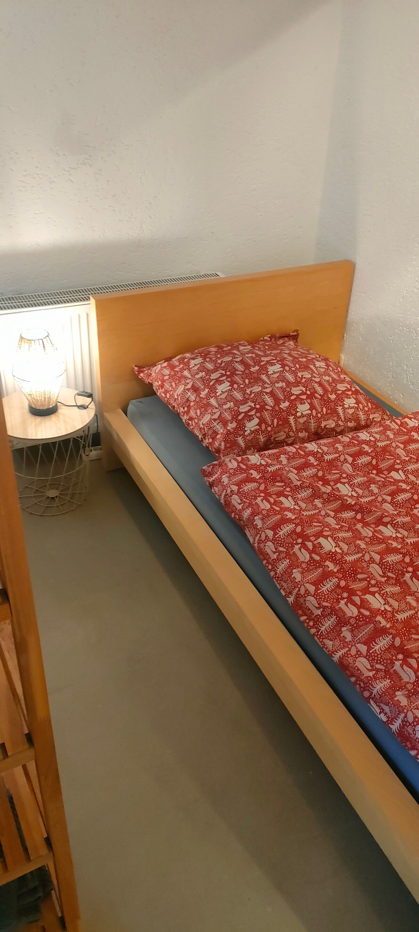 Une petite chambre avec un lit simple, une tête de lit en bois, une couvre-lit rouge et blanche à motifs et une lampe de chevet posée sur une table de chevet.