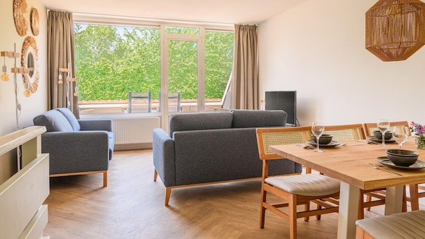Een moderne woonkamer met een bank, eettafel en stoelen. Er is een groot raam met gordijnen, een radiator en een hanglamp.