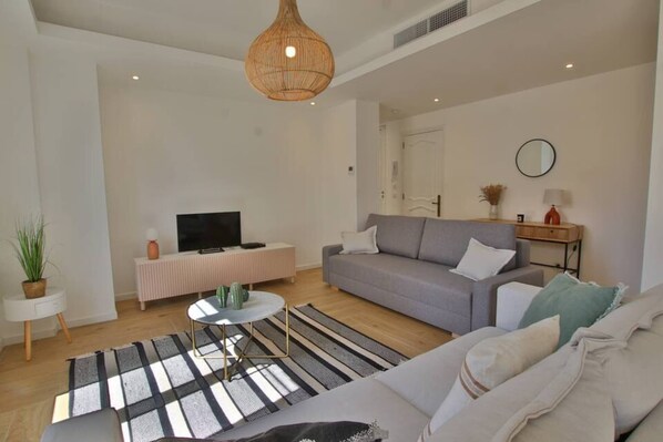 A modern living room with a grey sofa, a television on a white stand, a wicker pendant light, and a striped rug.