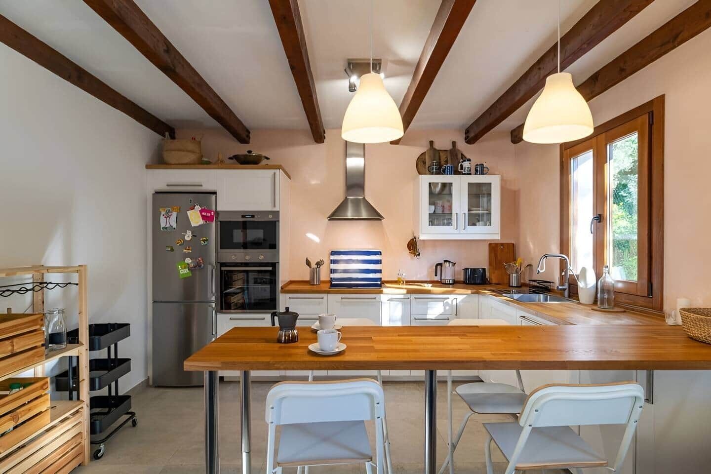 A modern kitchen with a central island, wooden beams, pendant lights, a refrigerator, oven, and a window providing natural light.