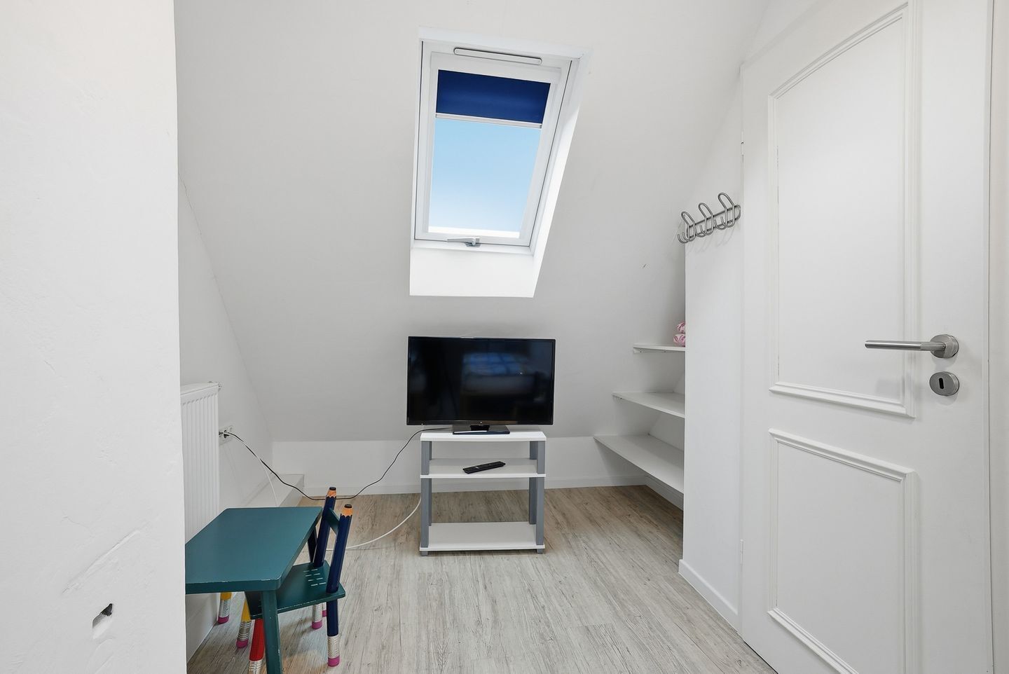 Una habitación con televisión sobre un pedestal, una mesita con sillas, una puerta y una ventana con vistas.