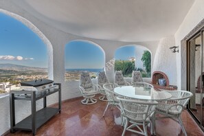 Un balcón con una parrilla, sillas de mimbre, una mesa de vidrio y vistas al paisaje.